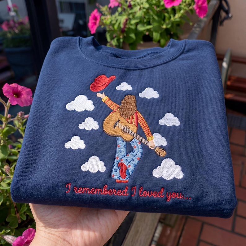 May include: A navy blue sweatshirt featuring an embroidered design of a person with long hair wearing a plaid shirt and star-patterned jeans, holding a guitar and a red cowboy hat, surrounded by white clouds and the text I remembered I loved you.
