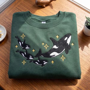 May include: A forest green crewneck sweatshirt featuring an embroidered design of four black and white orca whales swimming among several yellow four-pointed stars. The garment has a ribbed collar and long sleeves, with the embroidery centered.