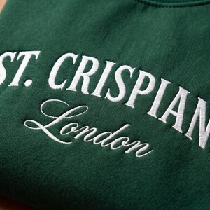 Może przedstawiać: Ciemnozielona bluza w kolorze lasu z białym haftowanym napisem ST. CRISPIANS pogrubioną czcionką szeryfową wielkimi literami, z wyrazem London napisanym poniżej białą kursywą w obszarze klatki piersiowej.