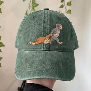 May include: A green baseball cap with an embroidered design of two monkeys. One monkey is pulling the other monkey by the arm. The cap is made of a soft, worn material.