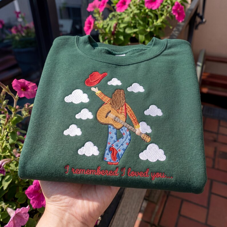 May include: A dark green crewneck sweatshirt featuring an embroidered design of a person with long hair holding a guitar and a red hat, surrounded by white clouds. The text "I remembered I loved you..." is embroidered in red below the figure.