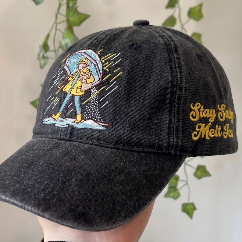 May include: Black baseball cap with an embroidered illustration of a person in a yellow raincoat and boots holding an umbrella. The text "Stay Salty Melt Ice" is embroidered in gold on the side. The cap is made of a washed, textured fabric.