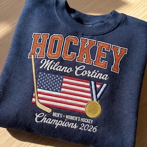 May include: Navy blue sweatshirt with embroidered text and graphics. The design includes the word "HOCKEY", "Milano Cortina", an American flag, a hockey stick, and a gold medal. The text "Champions 2026" is also featured.