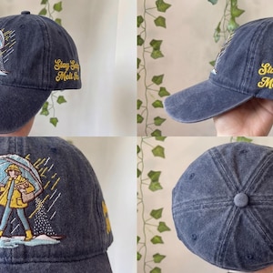 May include: A navy blue baseball cap with a curved brim. The cap features an embroidered illustration of a person in a yellow raincoat and boots holding an umbrella. The text "Stay Salty Melt Ice" is embroidered in yellow.