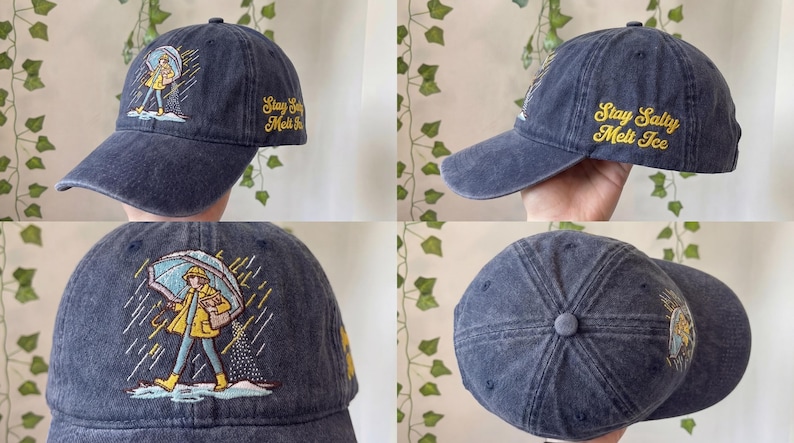 May include: A navy blue baseball cap with a curved brim. The cap features an embroidered illustration of a person in a yellow raincoat and boots holding an umbrella. The text "Stay Salty Melt Ice" is embroidered in yellow.
