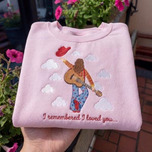 May include: A light pink crewneck sweatshirt featuring an embroidered design of a person with long brown hair wearing a plaid shirt and blue star-patterned pants, holding an acoustic guitar and a red hat, surrounded by white clouds and text.