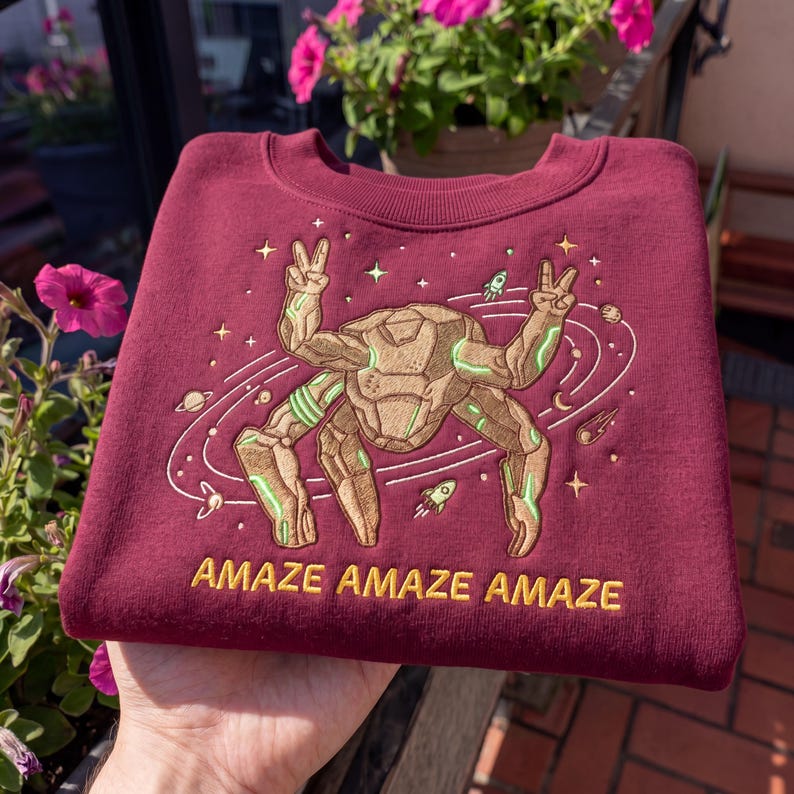 May include: A maroon crewneck sweatshirt featuring an embroidered design of a tan, multi-limbed rock creature making peace signs. The graphic includes white orbital rings, stars, planets, and rockets, with the text AMAZE AMAZE AMAZE in yellow.