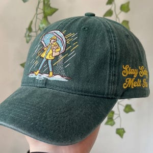 May include: Dark green baseball cap with an embroidered illustration of a person in a yellow raincoat and boots holding an umbrella. The text "Stay Salty Melt Snow" is embroidered in yellow on the side. The cap is made of a soft, textured fabric.