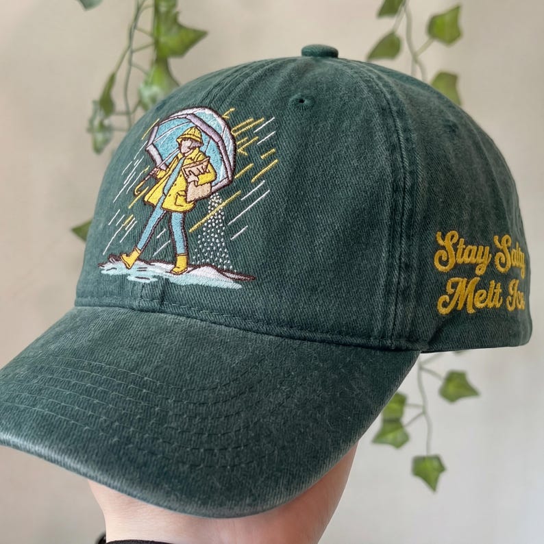 May include: Dark green baseball cap with an embroidered illustration of a person in a yellow raincoat and boots holding an umbrella. The text "Stay Salty Melt Snow" is embroidered in yellow on the side. The cap is made of a soft, textured fabric.