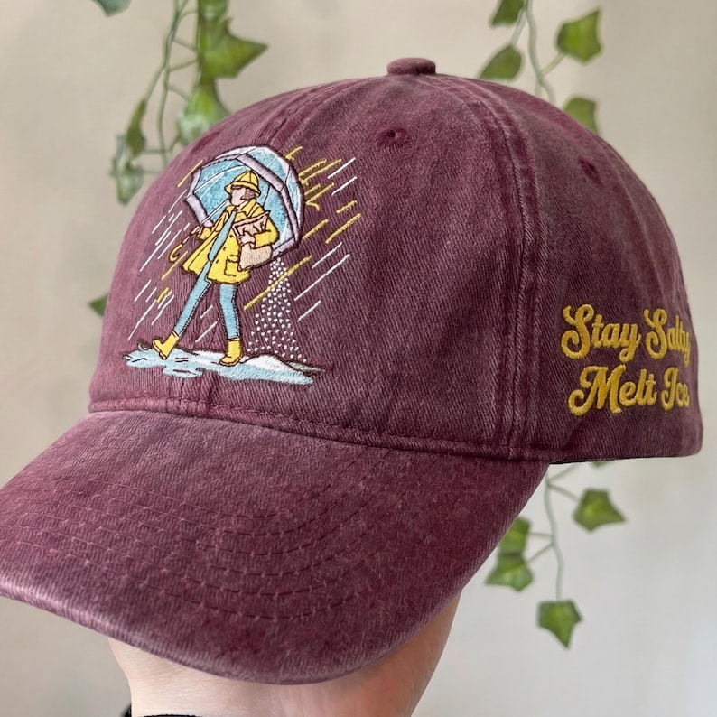 May include: A maroon baseball cap with an embroidered illustration of a person in a yellow raincoat and boots holding an umbrella. The text "Stay Salty Melt Ice" is embroidered in yellow on the side.