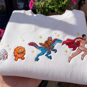 Pode incluir: Camisola branca com personagens de desenhos animados bordados. O design inclui uma mulher gritando, um monstro laranja, um gato montado em um dinossauro azul e um super-her&oacute;i correndo com uma capa vermelha. Estrelas douradas tamb&eacute;m s&atilde;o bordadas.