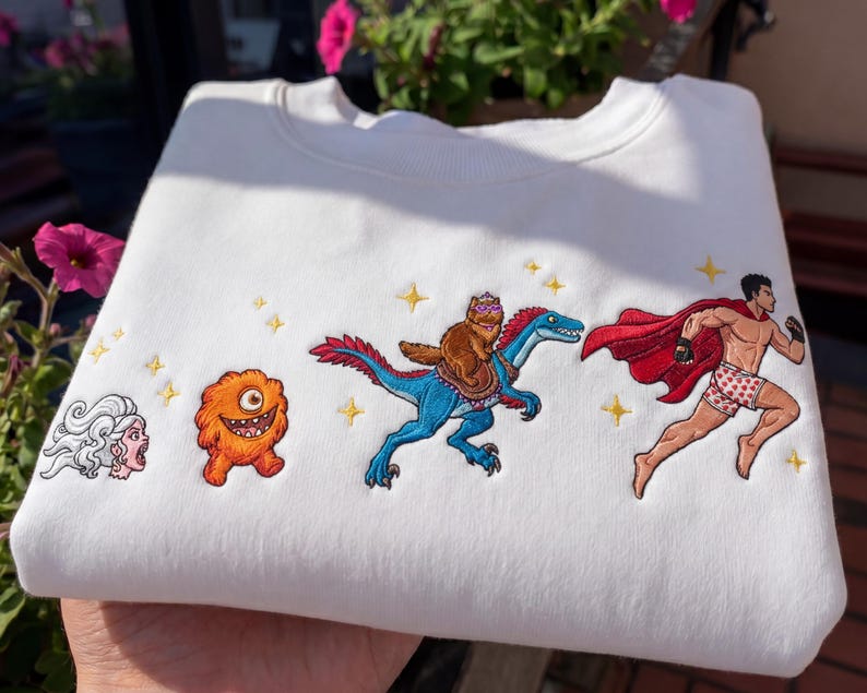 Pode incluir: Camisola branca com personagens de desenhos animados bordados. O design inclui uma mulher gritando, um monstro laranja, um gato montado em um dinossauro azul e um super-her&oacute;i correndo com uma capa vermelha. Estrelas douradas tamb&eacute;m s&atilde;o bordadas.