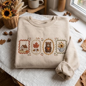 May include: A beige crewneck sweatshirt featuring four rectangular embroidered panels. The designs include a house with a pumpkin, a maple leaf with acorns, a brown bear holding a pumpkin, and a watering can filled with sunflowers.