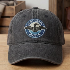 May include: A dark gray baseball cap with a circular patch featuring a loon design. The patch reads "Minnesota Anti Ice" in a blue and white color scheme. The cap has a curved brim and a fabric strap.