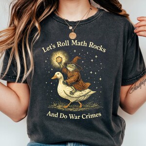 May include: Dark gray t-shirt with the text "Let's Roll Math Rocks And Do War Crimes." The graphic features a frog wizard riding a goose, holding a glowing die. The design is in shades of brown, yellow, and white.