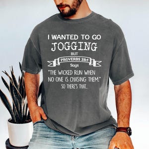 May include: Dark gray t-shirt with white text that reads "I WANTED TO GO JOGGING BUT PROVERBS 28:1 Says 'THE WICKED RUN WHEN NO ONE IS CHASING THEM.' SO THERE'S THAT..." The shirt is short-sleeved and has a relaxed fit.