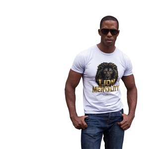 Peut inclure: T-shirt blanc avec un motif de lion noir et or et les mots "LION MENTALITY". Le t-shirt est associé à un jean bleu. La personne sur l'image porte des lunettes de soleil.