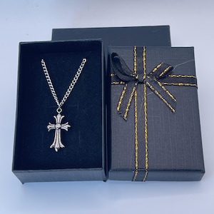 May include: A silver-colored cross pendant necklace displayed in an open black gift box. The necklace has a chain and a detailed cross. A second black gift box with a black bow and gold trim is next to it.