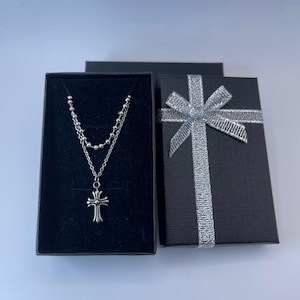 May include: A silver-colored necklace with a cross pendant, displayed in an open black jewelry box. A separate black gift box with a silver ribbon is next to it. The necklace has two strands of chain.