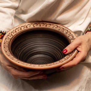 May include: A brown ceramic bowl with a decorative rim and a dark brown interior. The bowl is being held by a person's hands.