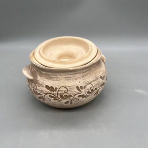 May include: A beige ceramic pot with a lid, featuring a floral design in a darker shade. The pot has two small handles and is suitable for kitchen use. The lid has a central opening. The overall design is rustic and handmade.