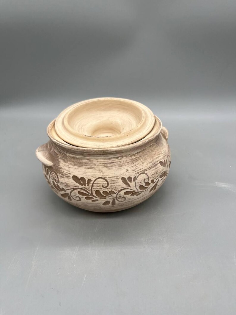May include: A beige ceramic pot with a lid, featuring a floral design in a darker shade. The pot has two small handles and is suitable for kitchen use. The lid has a central opening. The overall design is rustic and handmade.