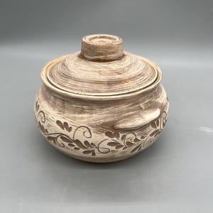 May include: A beige ceramic lidded bowl with a textured finish and floral design. The round bowl has a lid with a small handle and decorative handles on the sides. The floral design is etched into the ceramic.