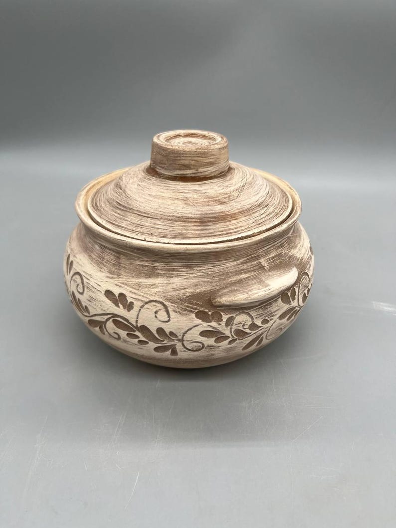 May include: A beige ceramic lidded bowl with a textured finish and floral design. The round bowl has a lid with a small handle and decorative handles on the sides. The floral design is etched into the ceramic.