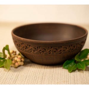 Op de afbeelding: Een bruine keramische kom met een ingewikkeld bloemmotief rond de rand. De kom staat op een neutrale ondergrond en wordt vergezeld door takjes met groene bladeren en kleine, beige bessen. De kom is leeg.