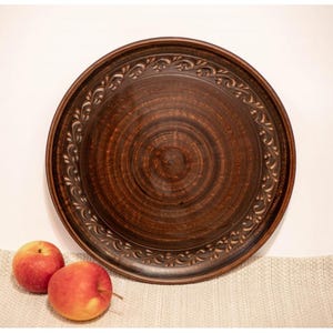 Może przedstawiać: Duży, okrągły, brązowy ceramiczny talerz z dekoracyjnym kwiatowym obramowaniem. Talerz ma spiralny wzór pośrodku i towarzyszą mu dwa czerwone jabłka. Talerz jest prawdopodobnie używany do serwowania potraw.