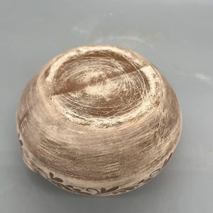 May include: A small, round, ceramic bowl with a textured, light brown exterior and a decorative floral pattern around the rim. The bowl's interior has a darker brown hue, and the overall design suggests a rustic or handmade aesthetic.