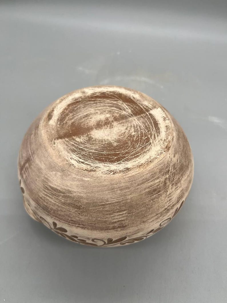 May include: A small, round, ceramic bowl with a textured, light brown exterior and a decorative floral pattern around the rim. The bowl's interior has a darker brown hue, and the overall design suggests a rustic or handmade aesthetic.