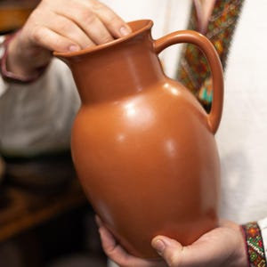 Puede incluir: Una jarra de cerámica de color terracota con un cuerpo redondeado y un asa curva. La jarra está sostenida por una persona que lleva una camisa blanca con detalles bordados coloridos. La jarra probablemente se utiliza para servir agua u otras bebidas.