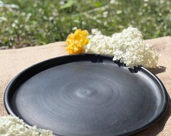 May include: A matte black ceramic plate with a wide rim. The plate is round and has a slightly textured surface. White and yellow flowers are arranged around the plate on a tan surface, with a green background.