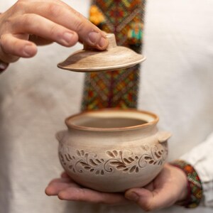 May include: A beige ceramic pot with a lid, held open to reveal the interior. The pot features a carved floral design around the base and small handles. The lid is also beige. The background is blurred.
