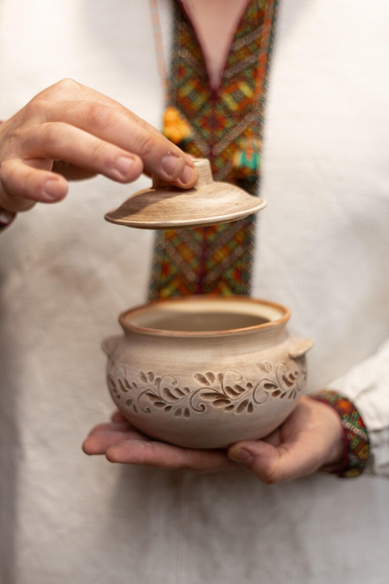 May include: A beige ceramic pot with a lid, held open to reveal the interior. The pot features a carved floral design around the base and small handles. The lid is also beige. The background is blurred.