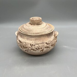 May include: A beige ceramic lidded pot with a decorative floral pattern around the base. The pot has two small handles and a knob on the lid. The pot is suitable for cooking and serving.