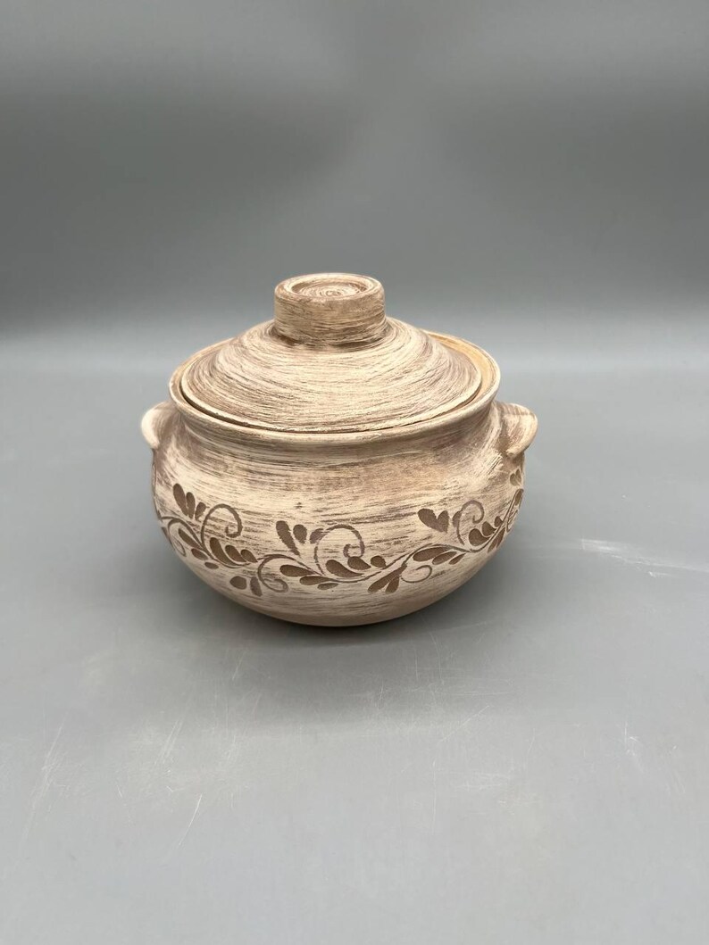 May include: A beige ceramic lidded pot with a decorative floral pattern around the base. The pot has two small handles and a knob on the lid. The pot is suitable for cooking and serving.