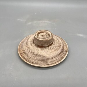 May include: A decorative, round, wooden object with a central, raised cylinder. The object has a light brown color with a textured, brushed finish. The item appears to be a lid or a small plate.