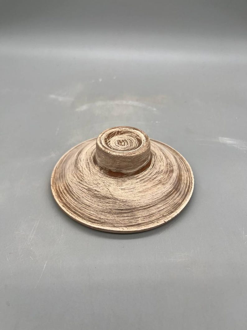 May include: A decorative, round, wooden object with a central, raised cylinder. The object has a light brown color with a textured, brushed finish. The item appears to be a lid or a small plate.