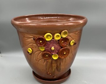 May include: A brown ceramic flower pot with a matching saucer. The pot features a raised floral design with yellow, maroon, and brown accents. The pot has a glossy finish and a wide opening.