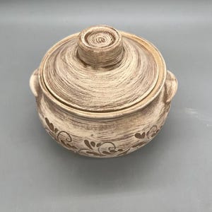 May include: A round, lidded ceramic pot with a textured, off-white and brown finish. The pot features two side handles and a decorative floral pattern around the base. The lid has a central knob.