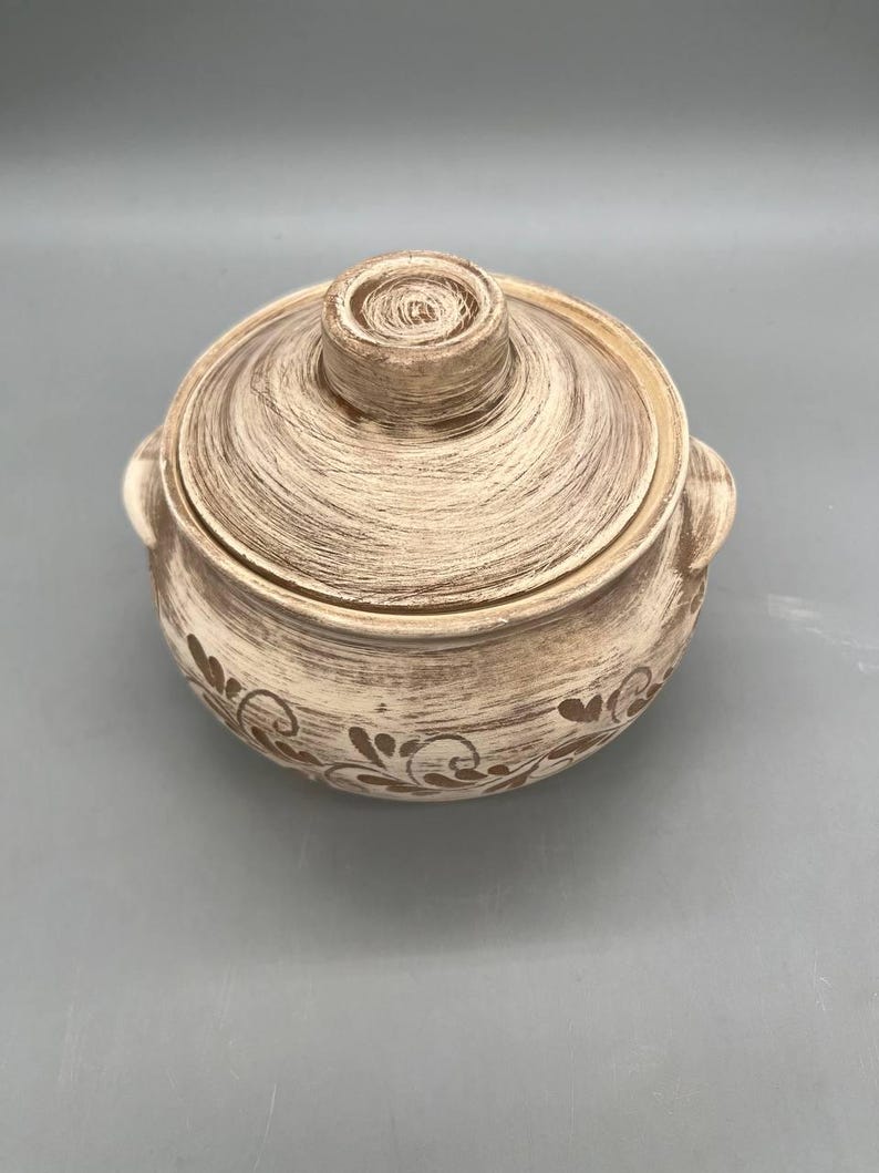 May include: A round, lidded ceramic pot with a textured, off-white and brown finish. The pot features two side handles and a decorative floral pattern around the base. The lid has a central knob.