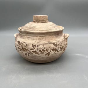 May include: A beige ceramic lidded pot with a decorative floral design. The pot has two small handles and a knob on the lid. The floral design is a darker shade of brown. The pot is likely used for cooking or serving.