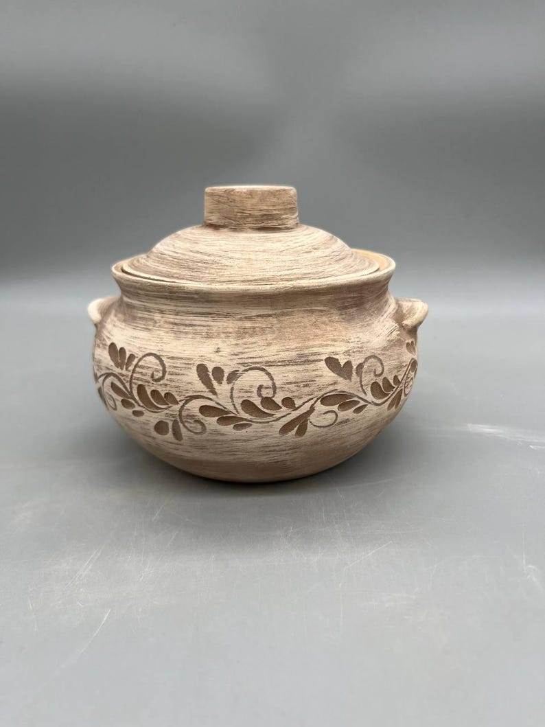 May include: A beige ceramic lidded pot with a decorative floral design. The pot has two small handles and a knob on the lid. The floral design is a darker shade of brown. The pot is likely used for cooking or serving.
