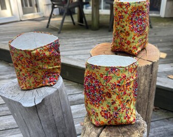 May include: Three fabric-covered containers with a floral pattern in red, orange, and yellow on a green background. The containers are placed on wooden stumps. The containers have a white interior.