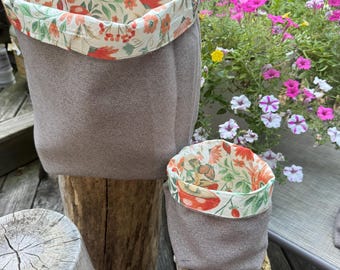 May include: Two fabric storage baskets in varying sizes. The exterior is a neutral brown, while the interior and top trim feature a floral pattern with orange, red, and green accents. The baskets are displayed on wooden stumps.