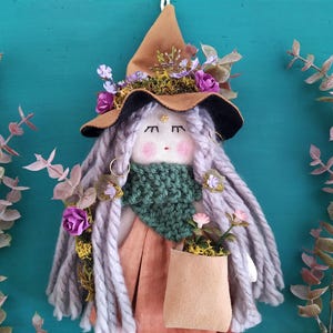 Puede incluir: Una muñeca hecha a mano con pelo largo de hilo gris, que lleva un sombrero de bruja de fieltro marrón con flores y una bufanda de punto verde. La muñeca lleva una bolsa de papel marrón.