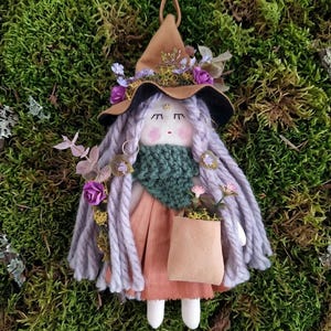 Puede incluir: Una muñeca de tela con un sombrero puntiagudo marrón adornado con flores moradas y azules, cabello largo de lana lavanda, bufanda de punto verde oscuro, vestido color melocotón y una pequeña bolsa beige, con rasgos faciales bordados y mejillas rosadas.