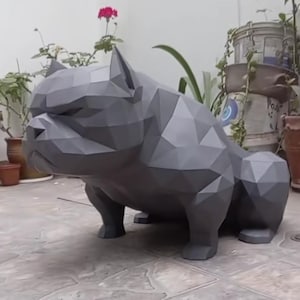 Könnte beinhalten: Eine graue, geometrische Papierkunst-Skulptur eines Bulldoggen in geduckter Position. Der Hund hat ein Low-Poly-Design, mit vielen flachen, abgewinkelten Oberflächen, die seine Form bilden. Die Skulptur steht auf einer gefliesten Oberfläche, mit Pflanzen und Töpfen im Hintergrund.